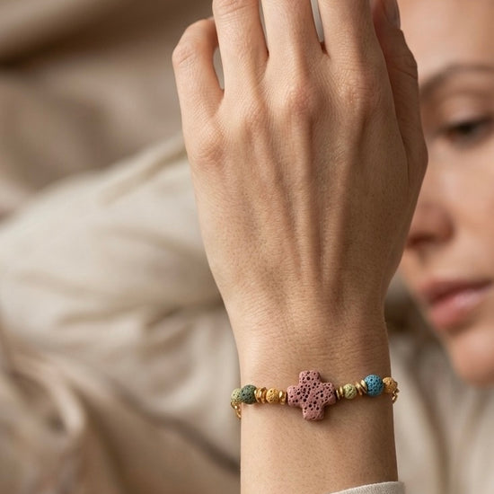 Pulsera con cruz de piedra volcánica en tonos multicolor, llevada en muñeca. Ideal para looks naturales y elegantes.