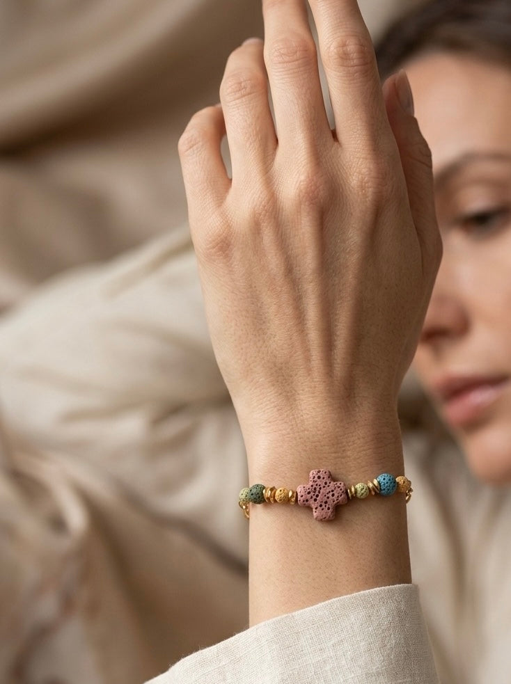 Pulsera con cruz de piedra volcánica en tonos multicolor, llevada en muñeca. Ideal para looks naturales y elegantes.